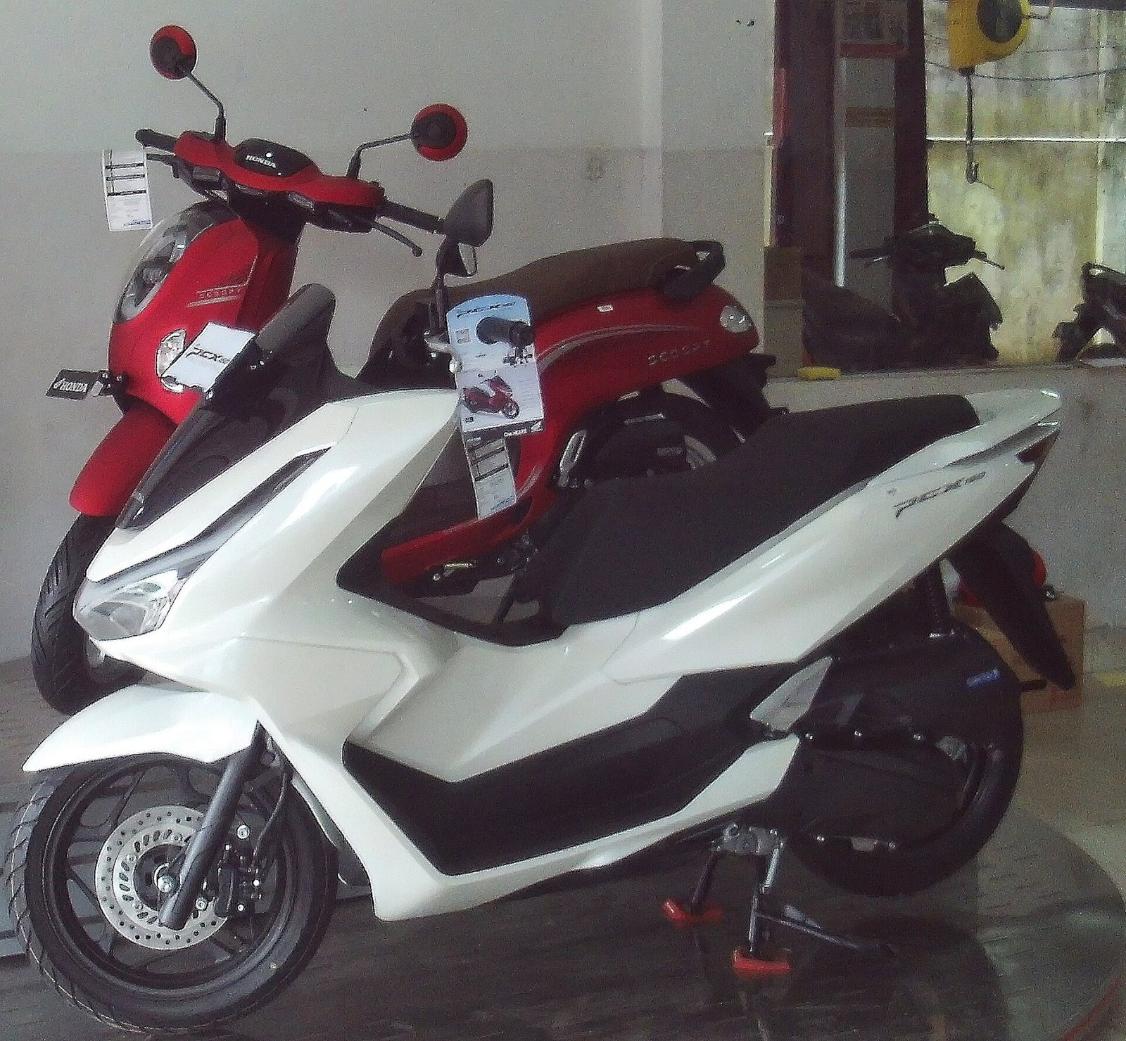 White Honda PCX in front of a red Honda Scoopy on a showroom floor