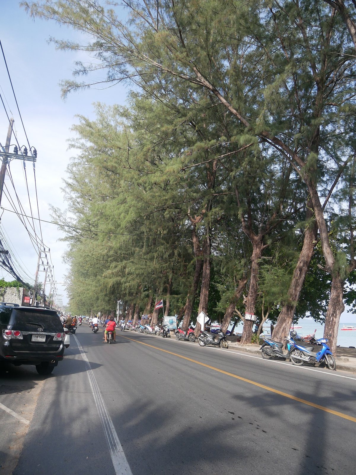 Tree lined coastal road at Rawai Beach in Phuket with traffic and parked motorbikes